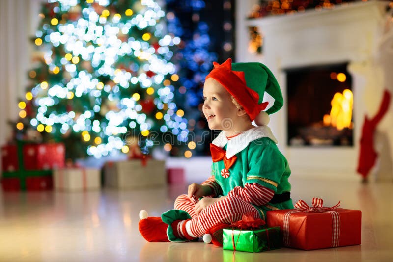 Child opening present at Christmas tree at home. Kid in elf costume with Xmas gifts and toys. Little baby boy with gift box and candy at fireplace. Family celebrating winter holidays. Home decoration. Child opening present at Christmas tree at home. Kid in elf costume with Xmas gifts and toys. Little baby boy with gift box and candy at fireplace. Family celebrating winter holidays. Home decoration