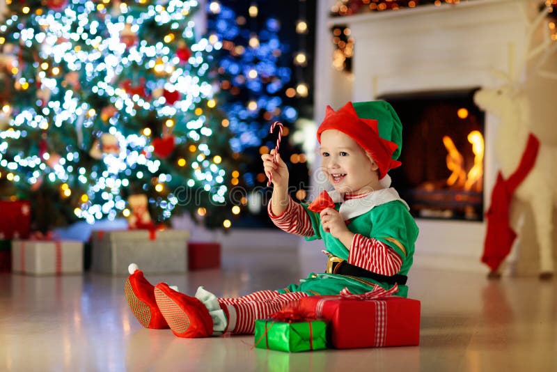 Child opening present at Christmas tree at home. Kid in elf costume with Xmas gifts and toys. Little baby boy with gift box and candy at fireplace. Family celebrating winter holidays. Home decoration. Child opening present at Christmas tree at home. Kid in elf costume with Xmas gifts and toys. Little baby boy with gift box and candy at fireplace. Family celebrating winter holidays. Home decoration