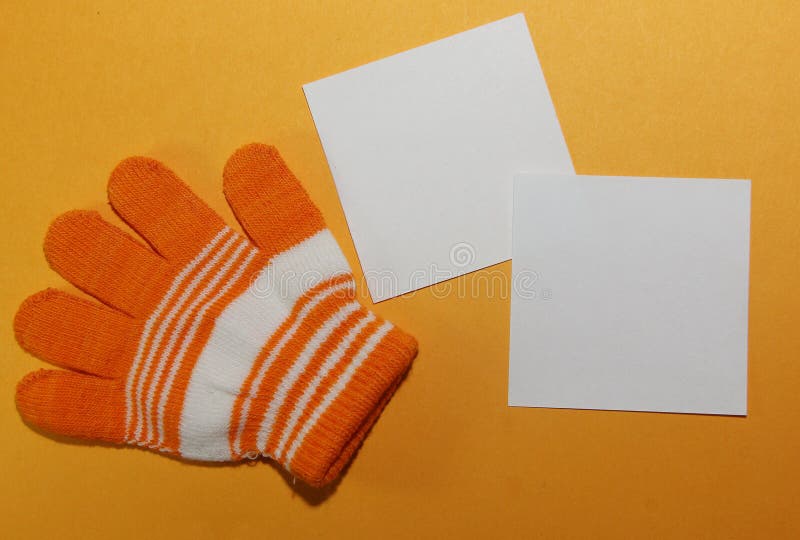 Kind- ` s Handschuh, auf der Kind-` s Hand, orange gestreifte Lügen auf einer gelben Oberfläche mit weißem Quadrat zwei formte Leerbelege für das Schreiben des Textes