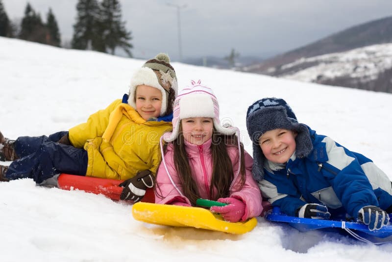 Three children, laying on their downhill sleds, stop to pose for this picture in the snow on a cold, wintry day. Three children, laying on their downhill sleds, stop to pose for this picture in the snow on a cold, wintry day.