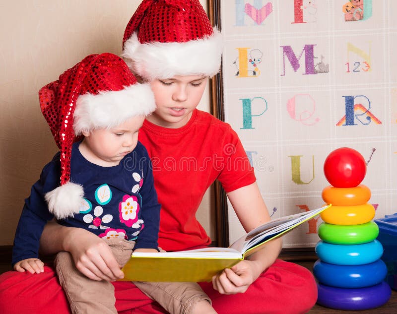 Kinderen die in Kerstmanhoeden een Kerstmisboek lezen Grote broerrea stock afbeeldingen Kinderen die in Kerstmanhoeden een Kerstmisboek lezen Grote broerrea stock afbeeldingen