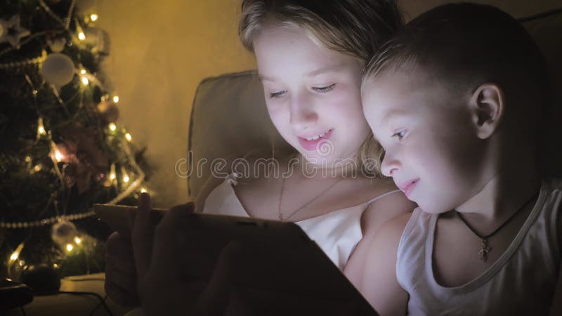 Kinderen spelen ' s nachts thuis tablet. twee kinderen spelen een slimme tablet op de achtergrond van de kerstboom.