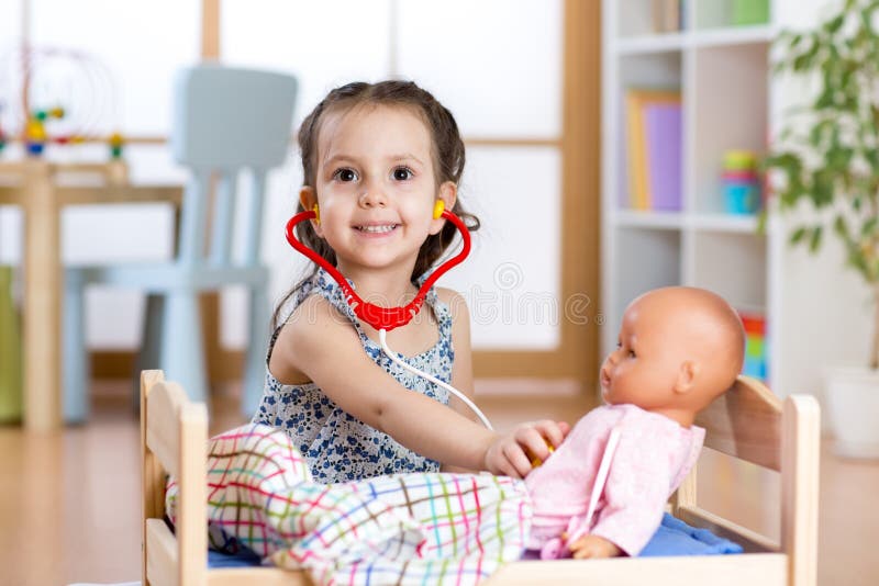 Kindermädchen, welches das Doktorrollenspiel überprüft ihre Puppe unter Verwendung des Stethoskops zu Hause sitzt im Spielzimmer