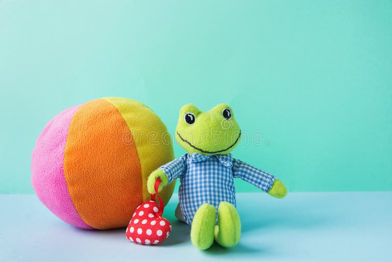 Kids Toys Small Plush Frog Holding Red Heart Multicolored Textile Soft Ball on Blue Green Background. Banner Placeholder Charity Nursery Hospital Copy Space. Kids Toys Small Plush Frog Holding Red Heart Multicolored Textile Soft Ball on Blue Green Background. Banner Placeholder Charity Nursery Hospital Copy Space