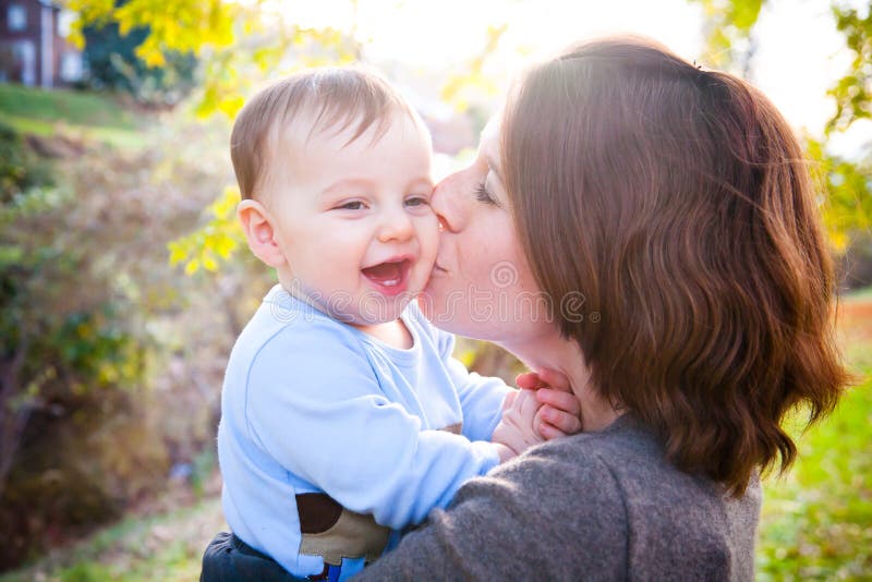 A mother kissing her young son on the cheek whose smile reveals two small teeth. The image is horizontal. A mother kissing her young son on the cheek whose smile reveals two small teeth. The image is horizontal.