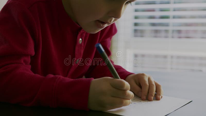 Kleine jongen in een rode trui die aan tafel zit, houdt hij een pen in zijn handen en schrijft of tekent op een wit blanco