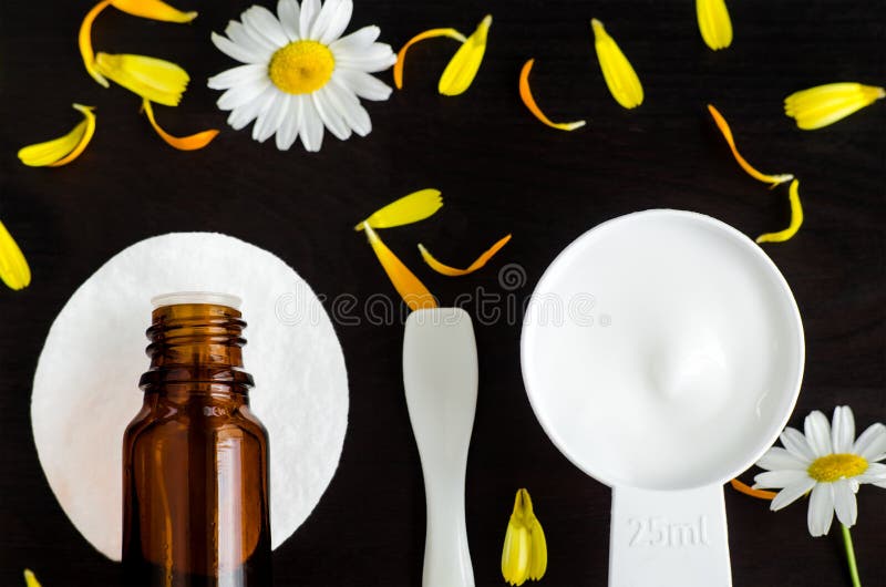 Small scoop with white mask/facial cream with flowers extracts. Bottle of essential oil. Chamomile, calendula marigold flowers dark background. Spa, natural skin care concept. Flat lay, copy space. Small scoop with white mask/facial cream with flowers extracts. Bottle of essential oil. Chamomile, calendula marigold flowers dark background. Spa, natural skin care concept. Flat lay, copy space.