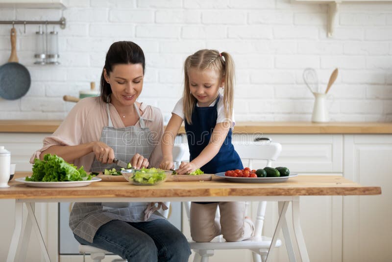 Kleine süße Tochter hilft jungen, stolzen Mama beim Schneiden von frischem Gemüse lizenzfreies stockfoto