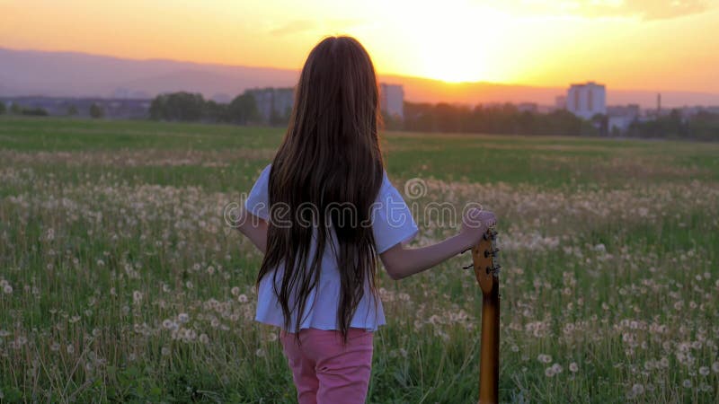 Am kleinen Musiker des Sonnenuntergangs hält Mädchen mitten in dem Feld Handgitarre und das Betrachten in den Abstand Stadt Konze