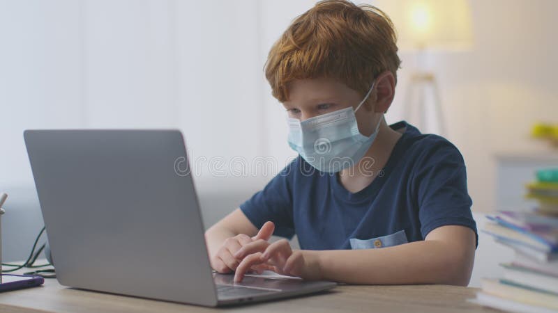Kleiner rothaariger Schuljunge mit schützender medizinischer Maske, die zu Hause online lernt und am Arbeitsplatz auf dem Laptop a stock video footage