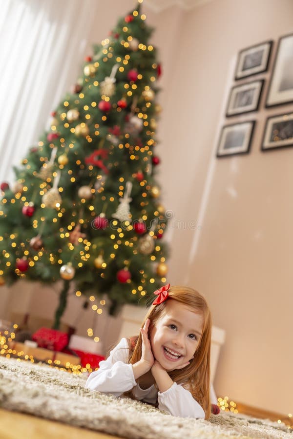 Kleines Mädchen, das auf dem Boden neben einem Weihnachtsbaum liegt. Ein schönes kleines Mädchen, das sich während eines Weihnachtsfestes zu Hause amüsiert und mit einem hübsch dekorierten Weihnachtsbaum und einem Lächeln auf dem Boden liegt