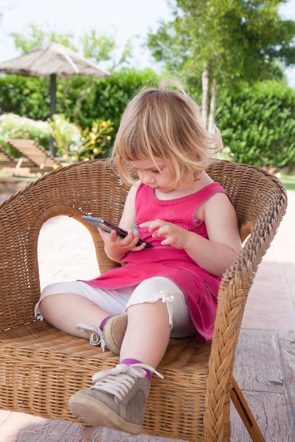 Kleines Mädchen, das an aufpassendem Handy des geflochtenen Stuhls sitzt lizenzfreies stockbild Kleines Mädchen, das an aufpassendem Handy des geflochtenen Stuhls sitzt lizenzfreies stockbild