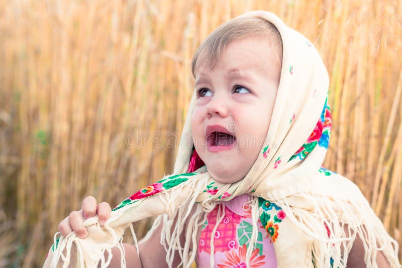Little girl in national scarf stands in the middle of the field and cries. Orphans, social problems. Little girl in national scarf stands in the middle of the field and cries. Orphans, social problems.