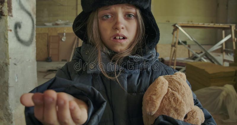Kleines obdachloses kaukasisches Mädchen in schmutzigen Winterbekleidung, das den Teddybären hält und um ein Handout bittet Hunge