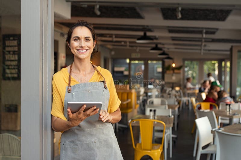 Portrait of happy woman standing at doorway of her store holding digital tablet. Cheerful mature waitress waiting for clients at coffee shop. Successful small business owner in casual clothing and grey apron standing at entrance and looking at camera. Portrait of happy woman standing at doorway of her store holding digital tablet. Cheerful mature waitress waiting for clients at coffee shop. Successful small business owner in casual clothing and grey apron standing at entrance and looking at camera