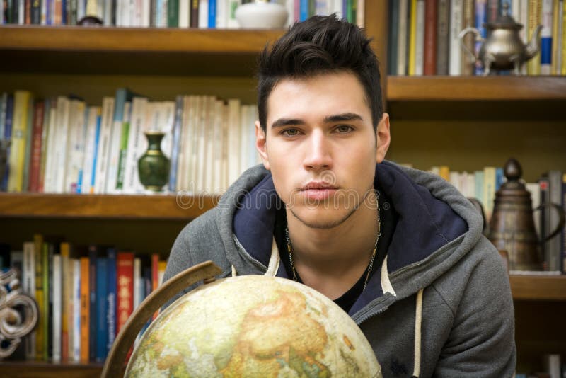 Handsome young man holding a globe indoors at home looking at the camera in front of a bookcase filled with books in a conceptual image. Handsome young man holding a globe indoors at home looking at the camera in front of a bookcase filled with books in a conceptual image