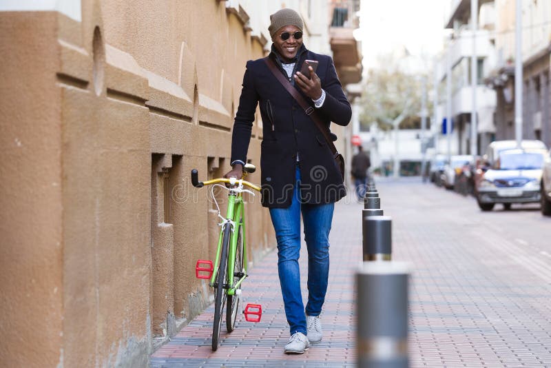 Knappe jonge mens die mobiele telefoon en staand vistuigfiets in de straat met behulp van royalty-vrije stock afbeelding Knappe jonge mens die mobiele telefoon en staand vistuigfiets in de straat met behulp van royalty-vrije stock afbeelding