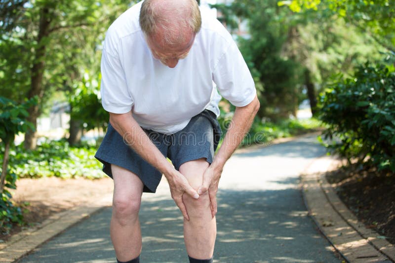 Closeup portrait, older man in white shirt, gray shorts, standing on paved road, in severe knee pain, isolated trees outside outdoors background.