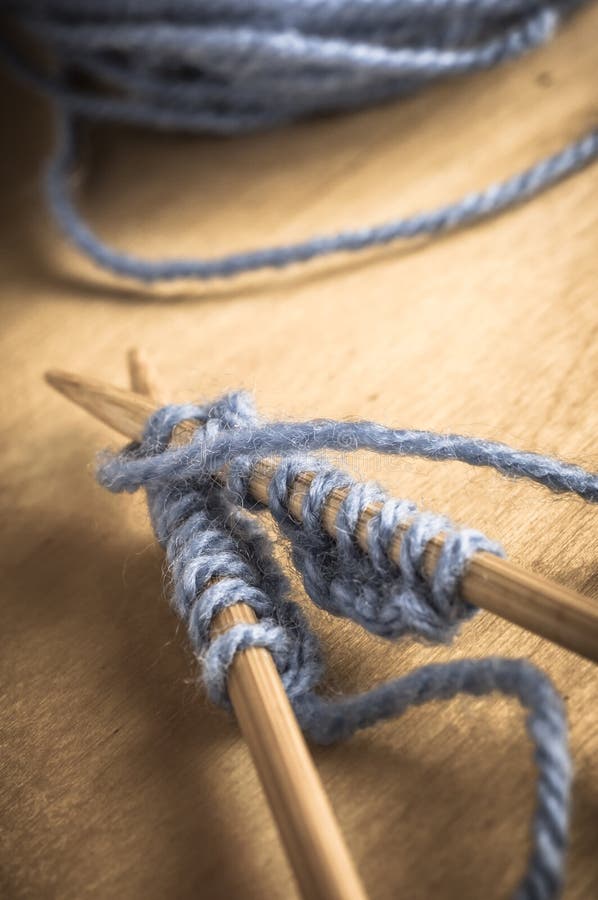 A crossed pair of knitting needles on wooden table containing stitches in progress with pale blue yarn and wool ball in the background. Slightly faded colours for vintage effect. A crossed pair of knitting needles on wooden table containing stitches in progress with pale blue yarn and wool ball in the background. Slightly faded colours for vintage effect.