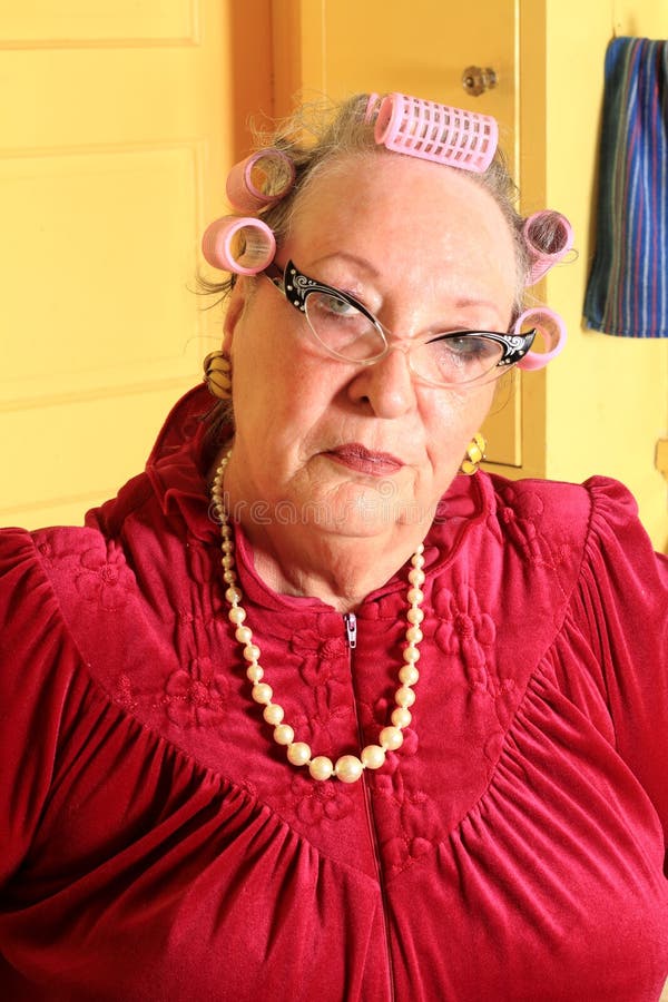 Silly Portrait of a grumpy senior gray haired granny lady wearing cat eye glasses, pearls and curlers in her hair in an old fashioned yellow kitchen. Silly Portrait of a grumpy senior gray haired granny lady wearing cat eye glasses, pearls and curlers in her hair in an old fashioned yellow kitchen.