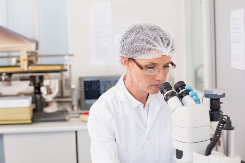 Kobieta naukowiec w płaszczu laboratoryjnym badająca próbkę pod mikroskopem na stanowisku laboratoryjnym z monitorem komputerowym zdjęcie stock