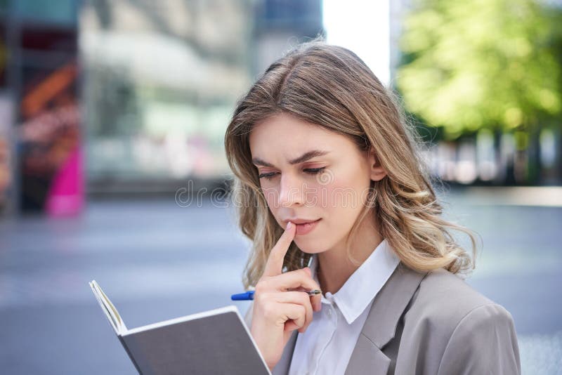 Woman looking concentrated while reading her notes, preparing for interview. Businesswoman in suit writes down in notebook, sits in city centre. Woman looking concentrated while reading her notes, preparing for interview. Businesswoman in suit writes down in notebook, sits in city centre.