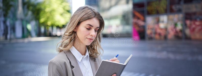 Woman looking concentrated while reading her notes, preparing for interview. Businesswoman in suit writes down in notebook, sits in city centre. Woman looking concentrated while reading her notes, preparing for interview. Businesswoman in suit writes down in notebook, sits in city centre.