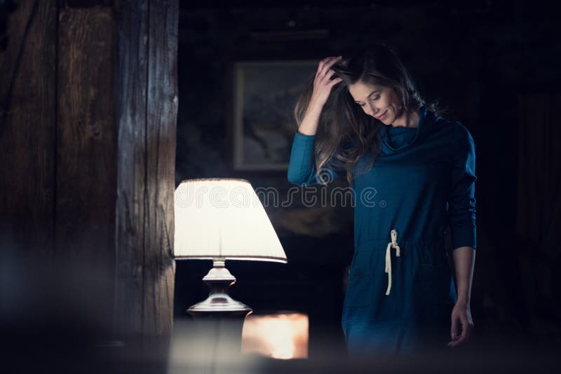Kobieta portret jest ubranym przypadkową suknię uśmiechniętą i patrzeje w dół blisko lampy je żyje pokój przy wiejski domowy salo fotografia stock Kobieta portret jest ubranym przypadkową suknię uśmiechniętą i patrzeje w dół blisko lampy je żyje pokój przy wiejski domowy salo fotografia stock