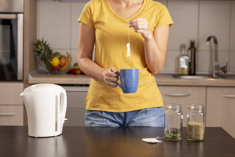 Woman making a cup of tea in the morning, holding a cup and adding a teabag into a boiling water from a kettle. Focus on the hand holding the cup. Woman making a cup of tea in the morning, holding a cup and adding a teabag into a boiling water from a kettle. Focus on the hand holding the cup