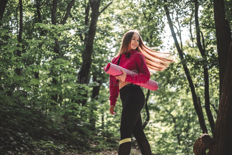 Kobieta trzyma sprawności fizycznej matę na trenować outdoors z trzepotliwym włosy w hoodie obrazy stock Kobieta trzyma sprawności fizycznej matę na trenować outdoors z trzepotliwym włosy w hoodie obrazy stock