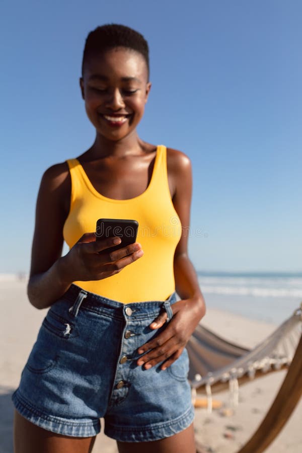 Kobieta używa telefon komórkowego z jej kciukami w pasku zapętla na plaży zdjęcia royalty free Kobieta używa telefon komórkowego z jej kciukami w pasku zapętla na plaży zdjęcia royalty free