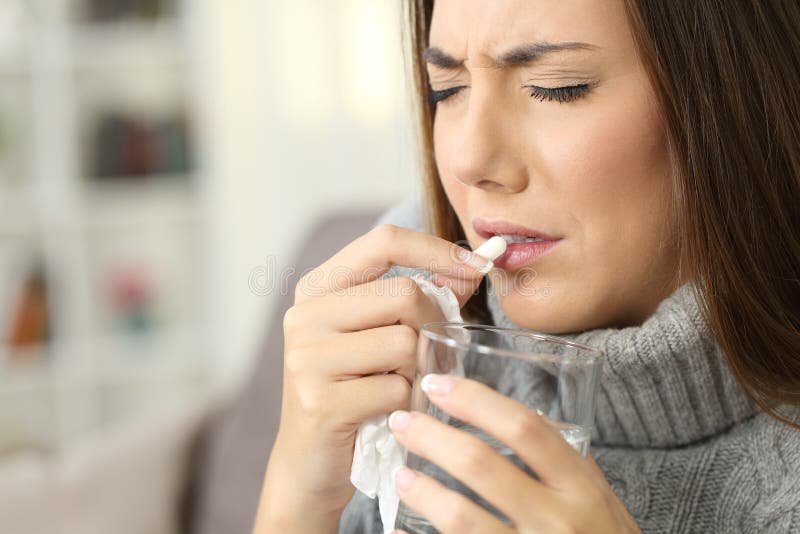 Sick woman wearing jersey suffering flu symptoms eating a pill sitting on a sofa in a house interior. Sick woman wearing jersey suffering flu symptoms eating a pill sitting on a sofa in a house interior