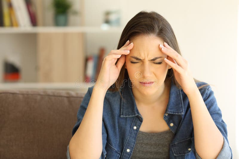 Front view portrait of a woman complaining suffering migraine sitting on a couch in the living room at home. Front view portrait of a woman complaining suffering migraine sitting on a couch in the living room at home