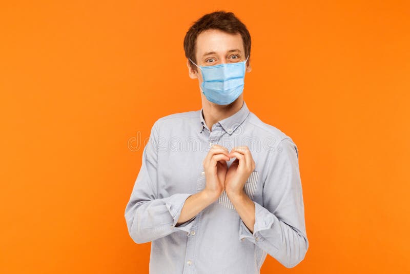 I love you. Portrait of joyful happy young worker man with surgical medical mask standing with heart love gesture and looking at camera with smile. indoor studio shot isolated on orange background. I love you. Portrait of joyful happy young worker man with surgical medical mask standing with heart love gesture and looking at camera with smile. indoor studio shot isolated on orange background
