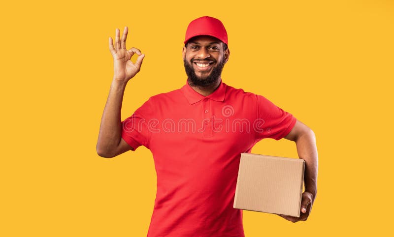 Fast Delivery. Black Courier Guy Holding Cardboard Box Gesturing Okay Posing On Yellow Studio Background. Fast Delivery. Black Courier Guy Holding Cardboard Box Gesturing Okay Posing On Yellow Studio Background