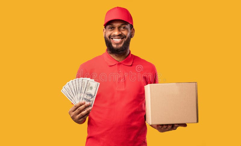 Pay On Delivery. African Courier Guy Holding Money Cash And Cardboard Box Standing On Yellow Background. Studio Shot. Pay On Delivery. African Courier Guy Holding Money Cash And Cardboard Box Standing On Yellow Background. Studio Shot