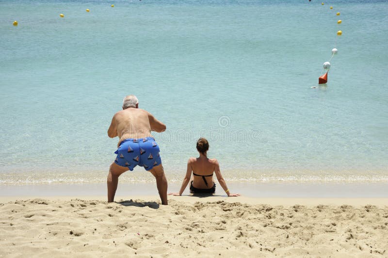 Komiskt och roligt läge En äldre man tar foto av den tillbaka sikten av den härliga flickan som sitter på stranden royaltyfri fotografi Komiskt och roligt läge En äldre man tar foto av den tillbaka sikten av den härliga flickan som sitter på stranden royaltyfri fotografi