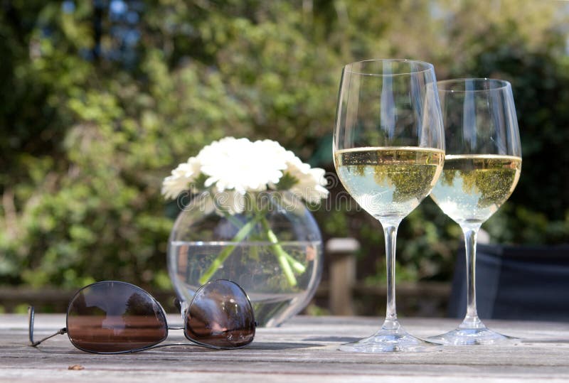 Two glasses of white wine, sunglasses, flowers on teak patio table. Two glasses of white wine, sunglasses, flowers on teak patio table