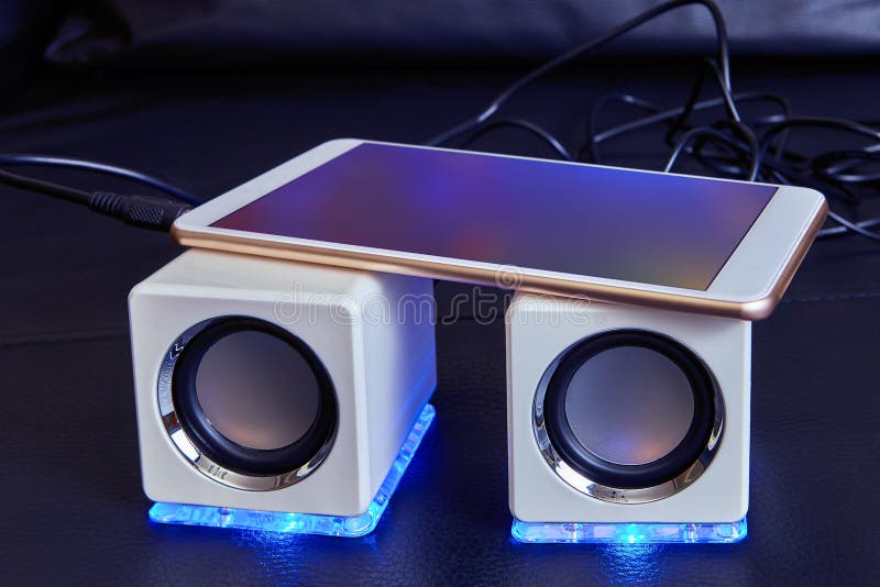 Two white mini speakers, of cubic form, with blue LED lights, are connected to the smartphone with a cable, the smartphone lies on top of the speakers. Two white mini speakers, of cubic form, with blue LED lights, are connected to the smartphone with a cable, the smartphone lies on top of the speakers