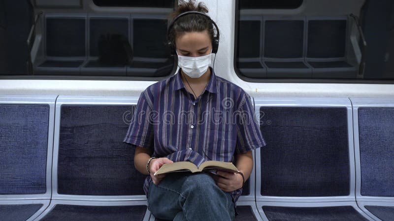 Koncentrerad maskerad årtusende som läser en bok och lyssnar på musik på tunnelbana stock video