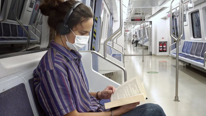 Koncentrerad maskerad årtusende som läser en bok och lyssnar på musik på tunnelbana stock video