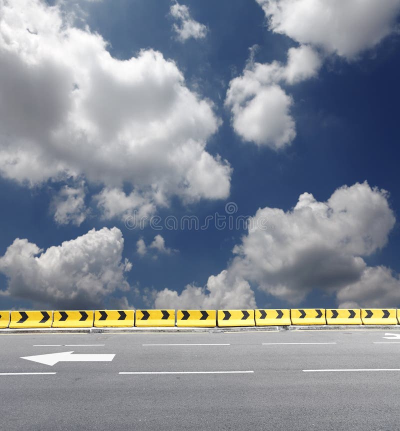 Rows of yellow concrete safety barrier with directional arrow on a road kerb against a blue cloudy sky. Rows of yellow concrete safety barrier with directional arrow on a road kerb against a blue cloudy sky.