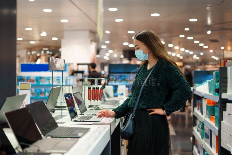 Konzentrierte junge Frau in Gesichtsmaske untersucht Laptop im Technikfachgeschäft stockbild