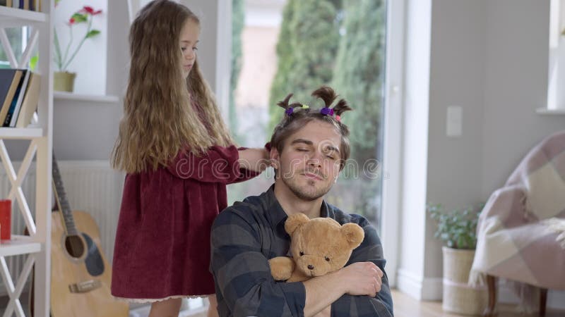 Konzentriertes kleines Mädchen, das Pferdeschwänze auf dem Haar des freudigen Vaters macht und Eltern Ohren zieht. positiv freudig stock video footage