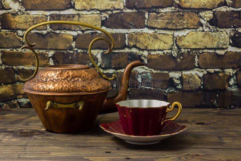 Old copper kettle with contrast of magenta bone china cup, on a wooden table with exposed brick wall. Old copper kettle with contrast of magenta bone china cup, on a wooden table with exposed brick wall