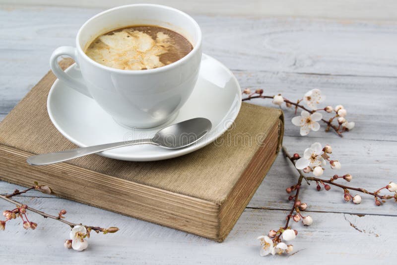 Kopp kaffe med kräm på den gamla boken, träbakgrund med blommande körsbärsröda filialer royaltyfri fotografi Kopp kaffe med kräm på den gamla boken, träbakgrund med blommande körsbärsröda filialer royaltyfri fotografi