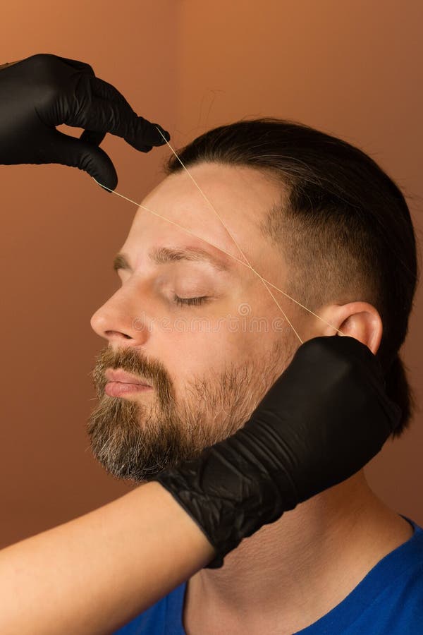 Brutal male with beard gets brow correction procedure. Barber doing threading and correcting shape of eyebrows to male client in barber shop. Brow care concept. Brutal male with beard gets brow correction procedure. Barber doing threading and correcting shape of eyebrows to male client in barber shop. Brow care concept.