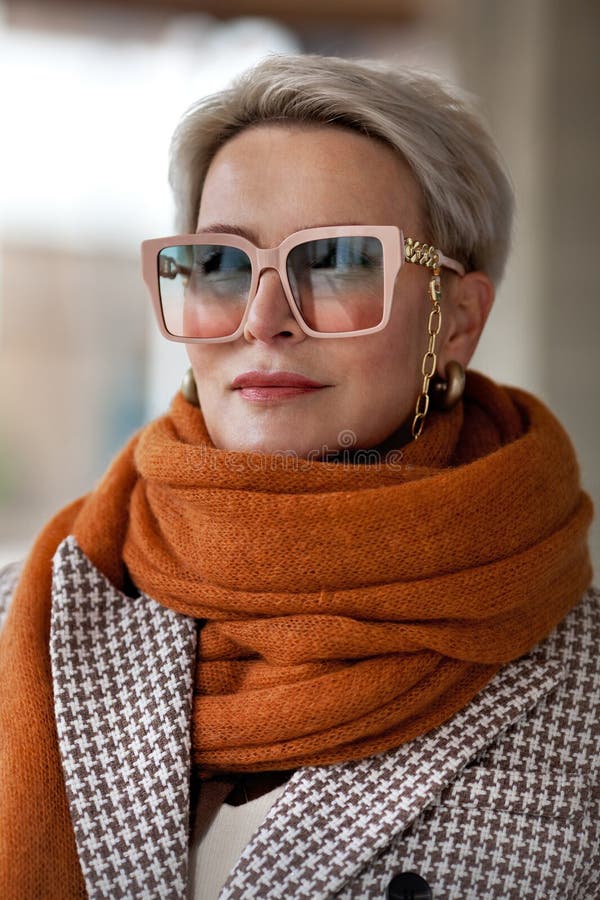 Portrait woman short blonde hair in warm knitted ochre scarf, double-breasted jacket and fashionable glasses. Fashion style autumn or spring, female fashionista 50 years old. Portrait woman short blonde hair in warm knitted ochre scarf, double-breasted jacket and fashionable glasses. Fashion style autumn or spring, female fashionista 50 years old.