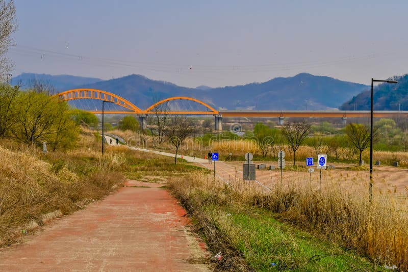 Sejong, South Korea; April 1, 2020: Landscape of public park with unrecognizable people riding bicycles and yellow truss bridge in background. Sejong, South Korea; April 1, 2020: Landscape of public park with unrecognizable people riding bicycles and yellow truss bridge in background