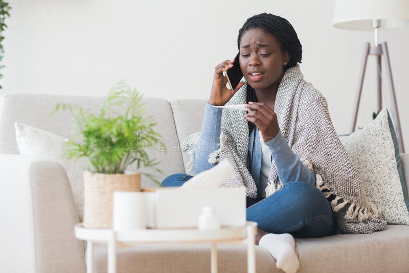 Sick african american girl sitting on couch, holding thermometer and calling family doctor for counseling, free space. Sick african american girl sitting on couch, holding thermometer and calling family doctor for counseling, free space